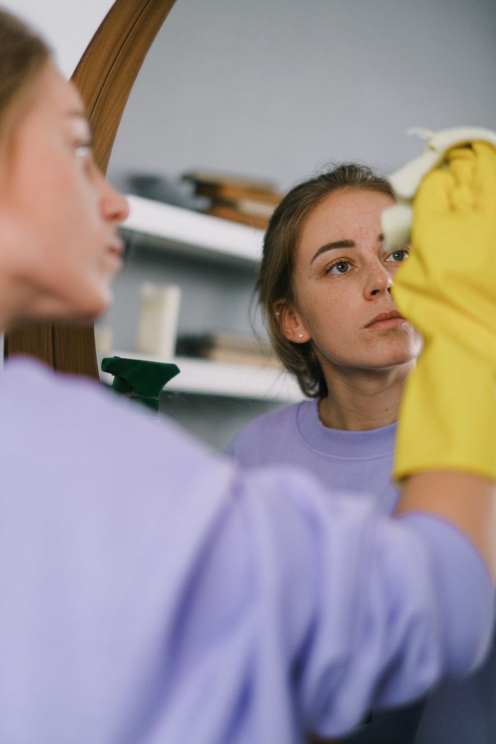 Crop female in purple sweatshirt and latex gloves washing mirror in light apartment in daytime